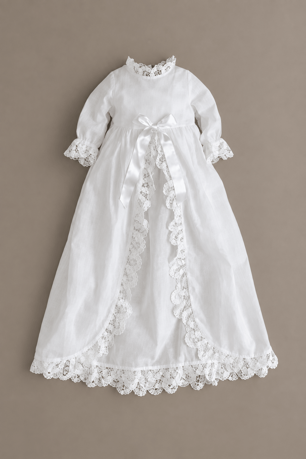 A short, white long-sleeve baby or toddler christening dress with delicate lace trim, a high lace collar, and a satin bow at the waist, displayed flat against a solid taupe background.