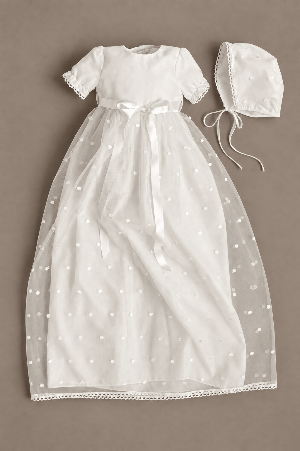 A white baby christening dress with short sleeves, a sheer tulle skirt with embroidered dots, and a matching bonnet, laid flat on a neutral taupe background.