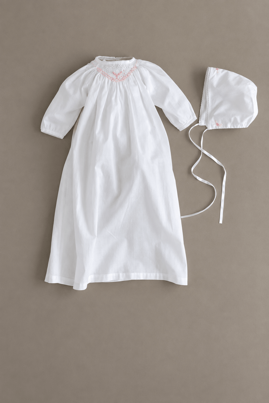 A white baby baptism gown with three-quarter sleeves and delicate pink embroidery around the neckline, shown next to a matching white bonnet with ties on a solid taupe background.