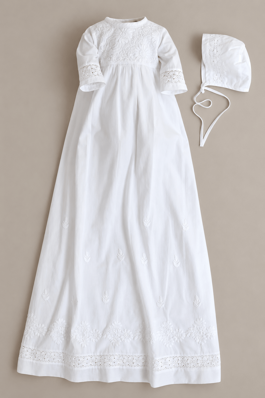 A white long christening dress with short sleeves, lace trim at the cuffs and hem, and a matching bonnet with a tie, laid flat on a neutral beige background.