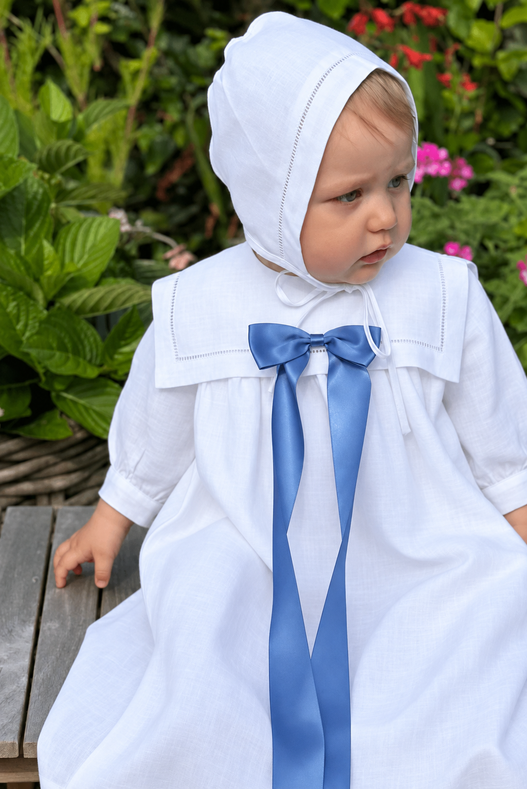 A toddler wearing a white monterey christening dress with a wide collar and a long blue ribbon bow at the chest, seated outdoors on a wooden bench with green garden foliage in the background.
