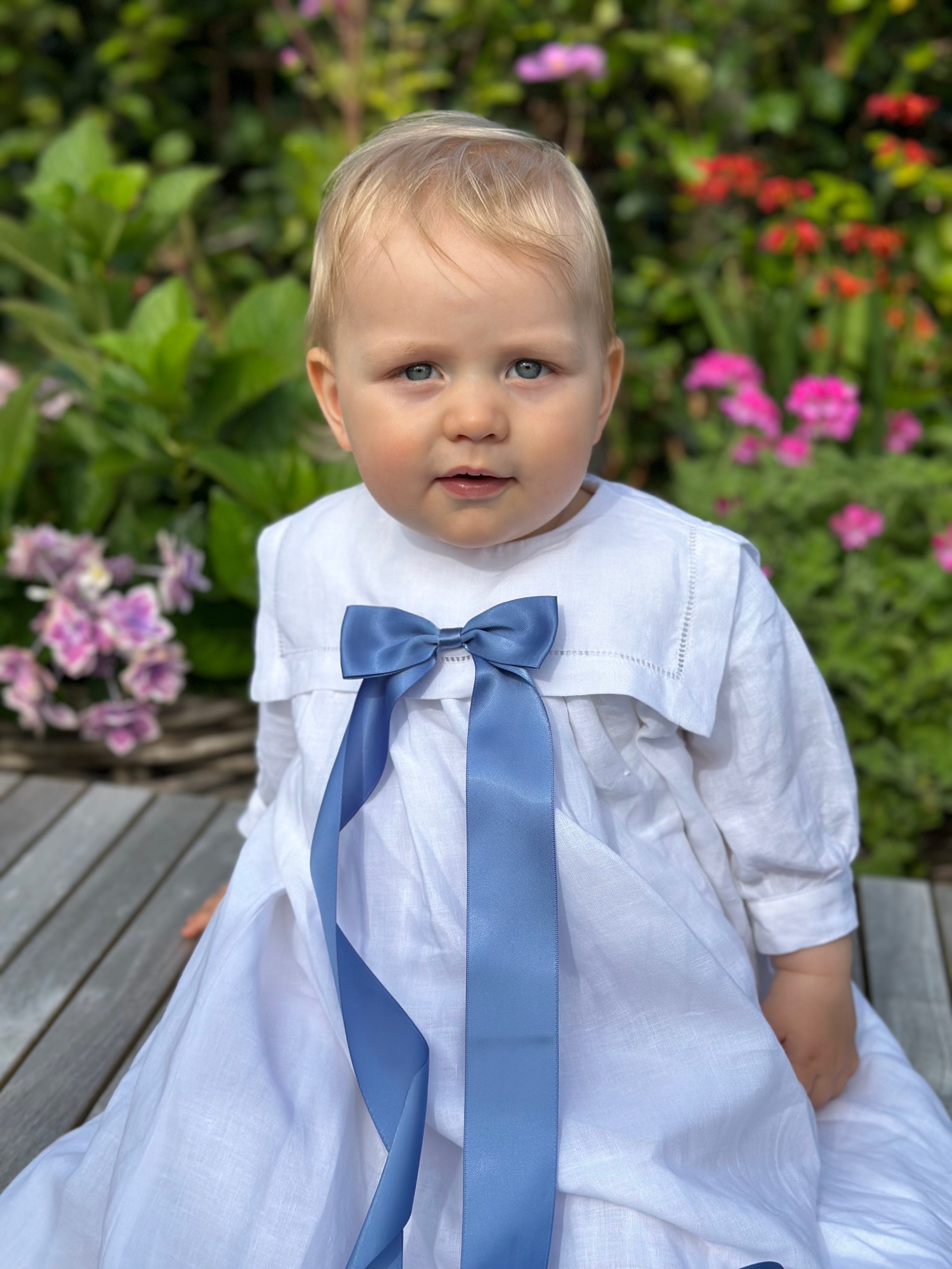 Child wearing a white christening gown with a blue ribbon in a garden setting