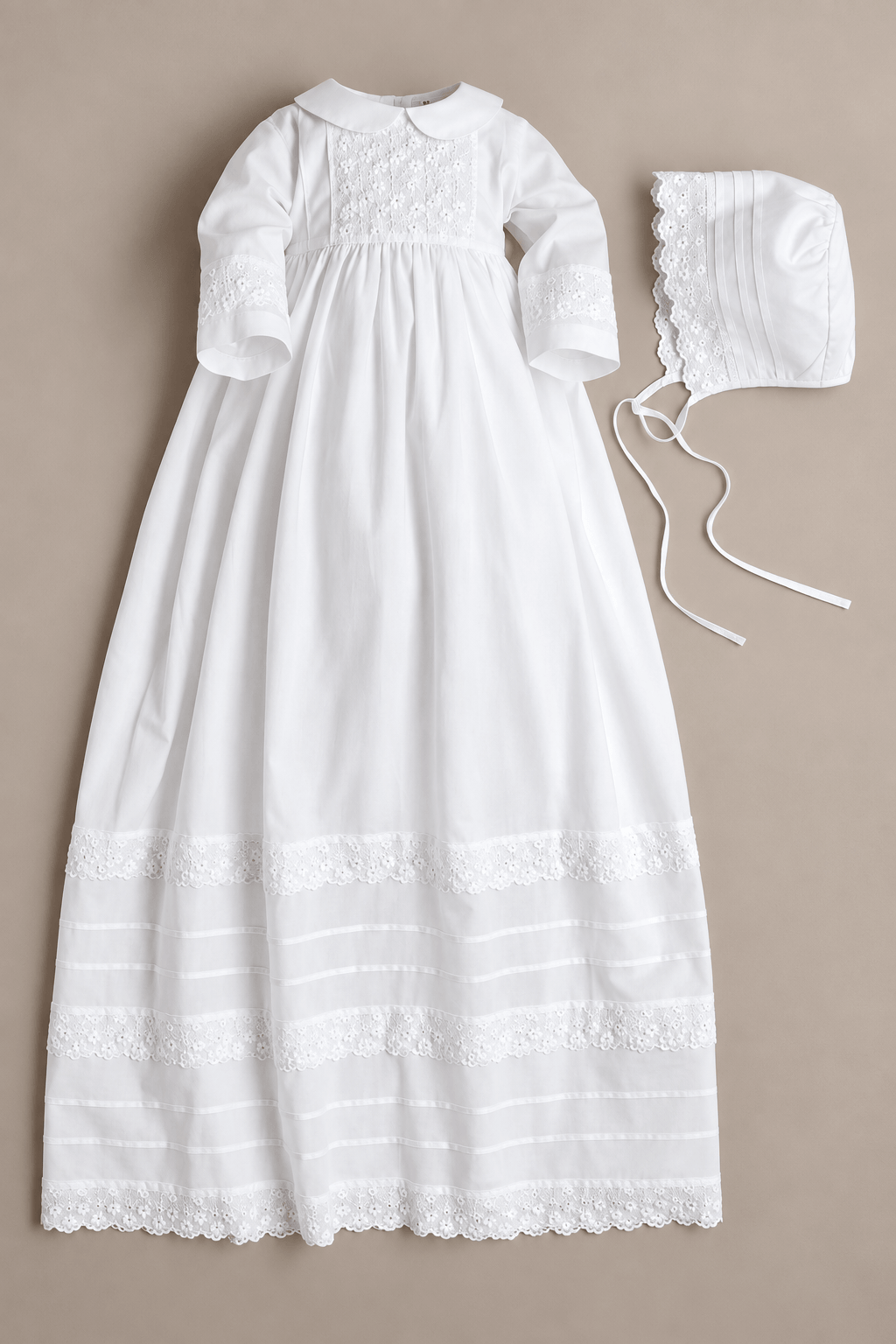 A white long-sleeve baby christening dress with embroidered detailing and lace trim, shown flat-laid next to a matching white bonnet on a solid beige background.