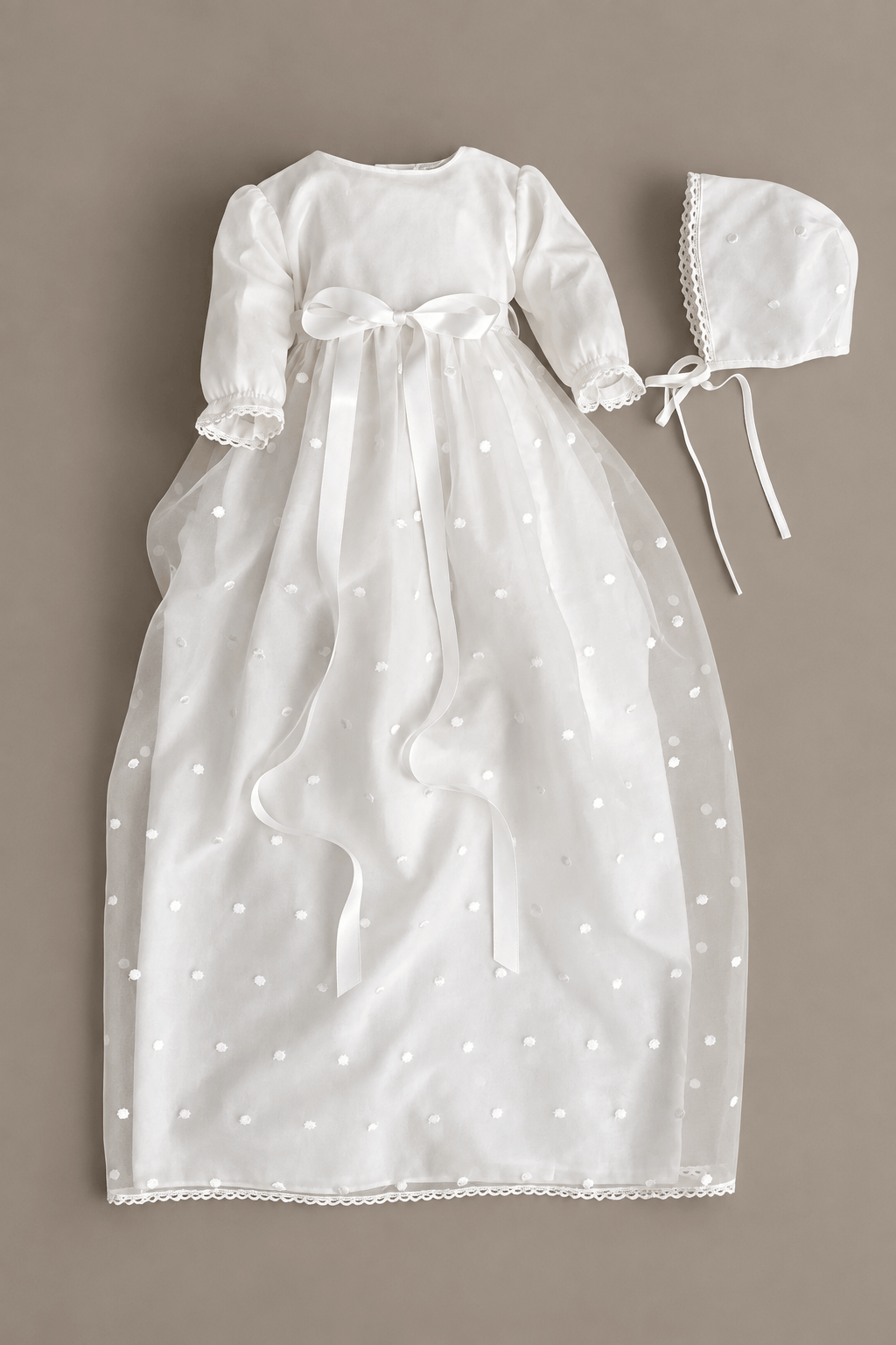 A off-white baby girl's christening dress with long sleeves, a bow at the waist, and a matching bonnet, laid flat on a neutral taupe background.