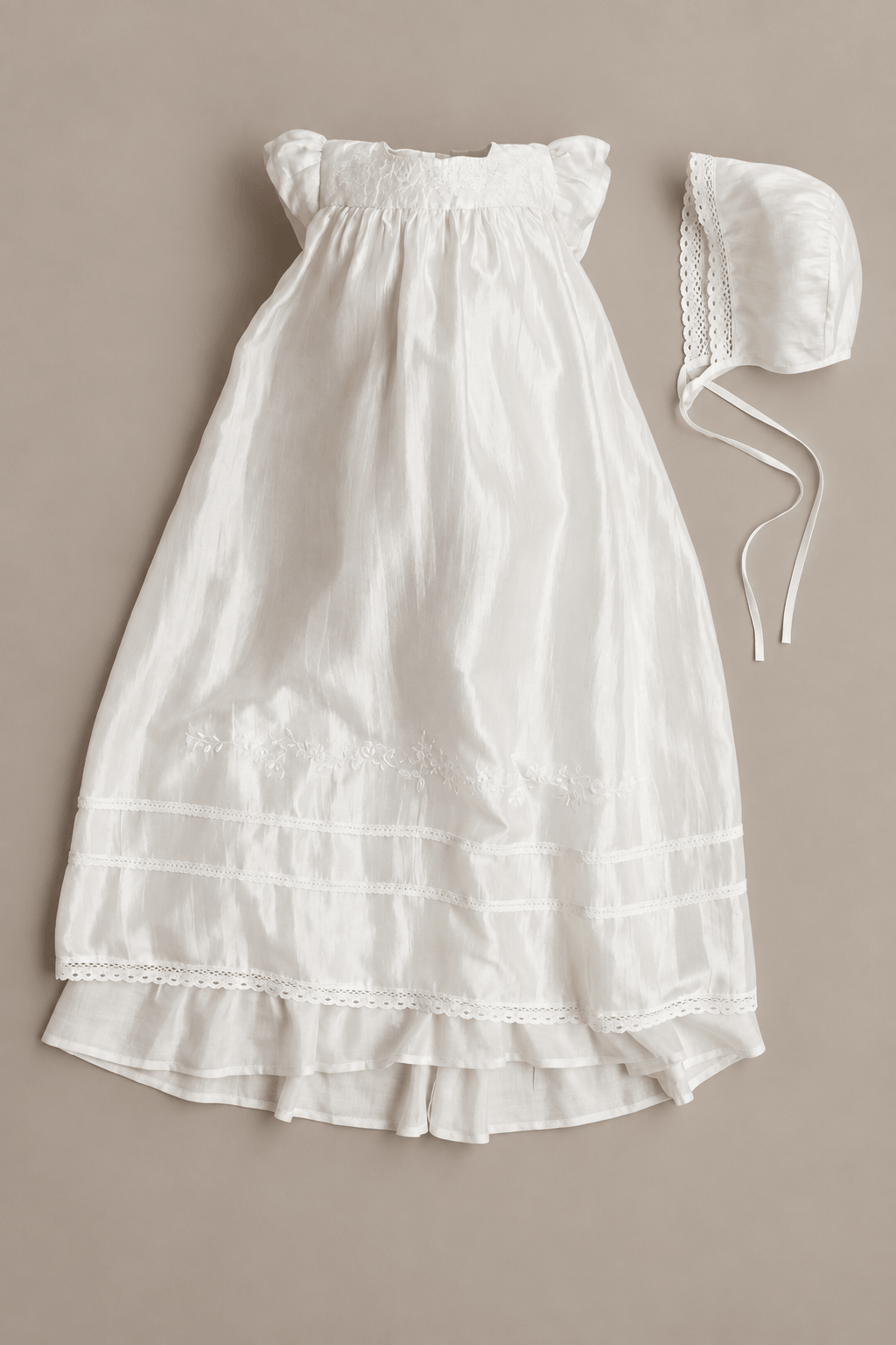 A white baby christening dress with a smocked bodice and ruffled hem, shown flat-laid next to a matching white bonnet on a solid taupe background.