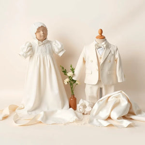 White christening gown and light tailored suit styled with bonnet and satin accessories on a neutral backdrop