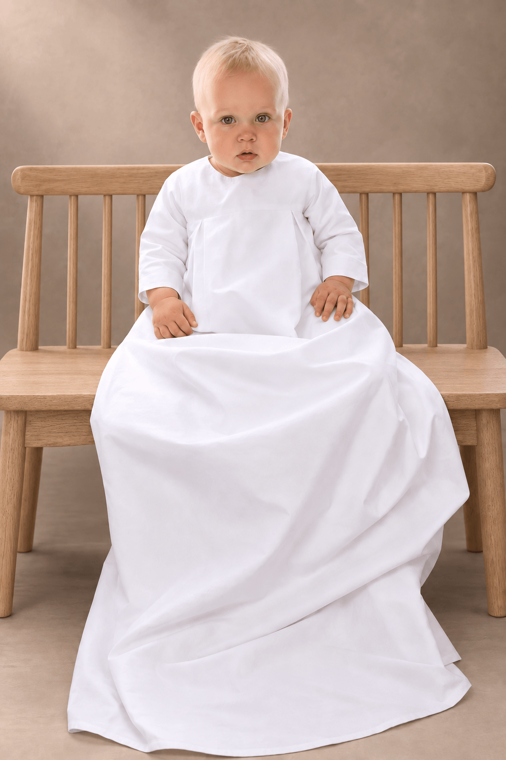 A baby with light hair sits on a wooden bench wearing a white long-sleeve christening gown set against a neutral studio background.