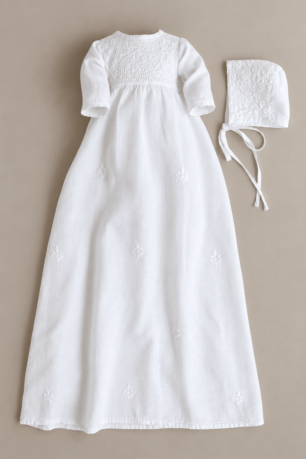 A white baby christening dress with short sleeves and embroidered details, shown flat-laid next to a matching white bonnet on a neutral beige background.