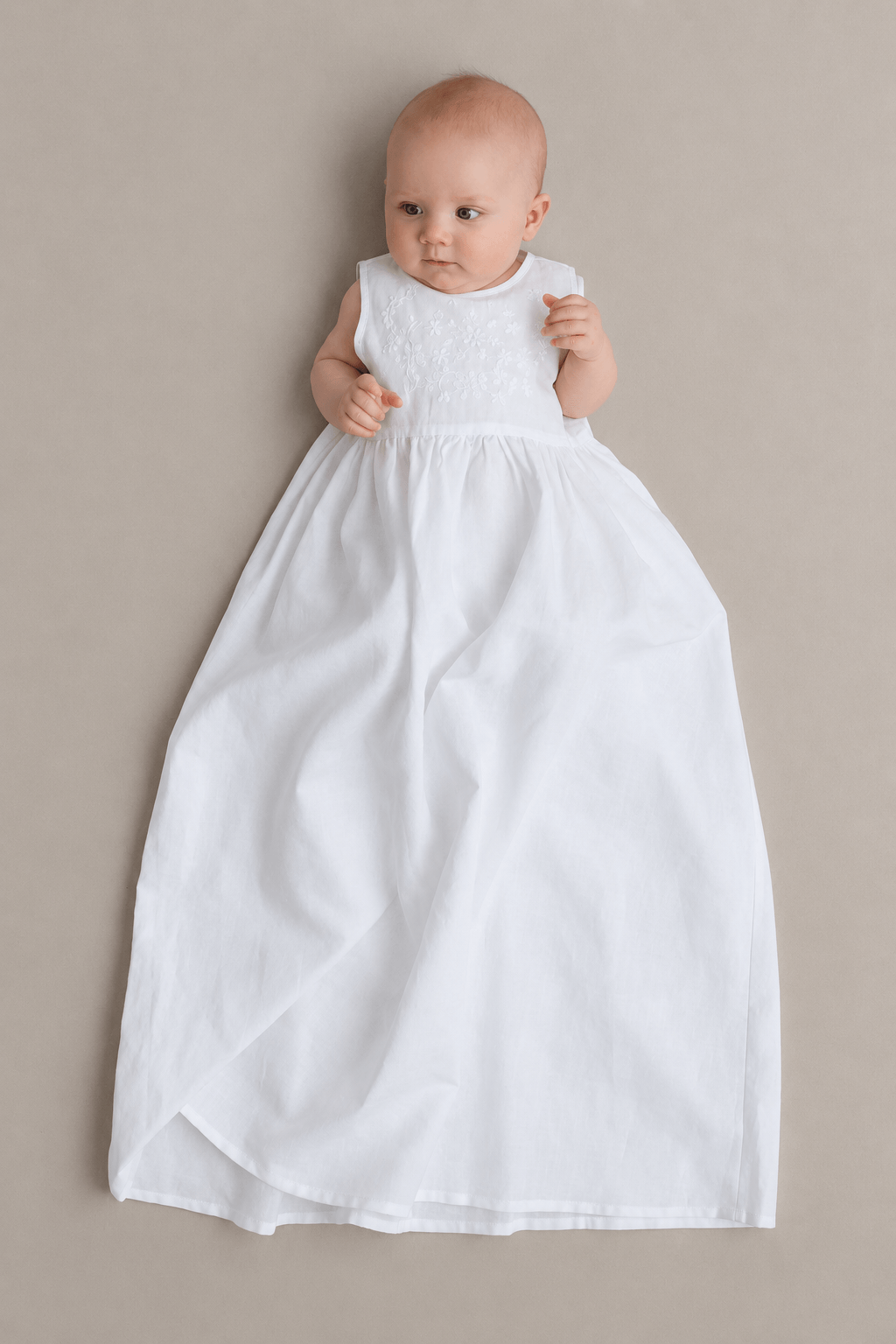 A baby wearing a white sleeveless christening dress with a textured yoke and a full skirt, lying on a neutral beige background.