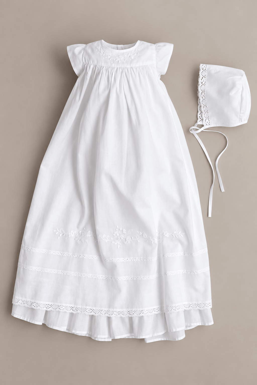 A white baby girl's baptism dress with short flutter sleeves, a smocked bodice, and lace trim, shown with a matching white bonnet on a neutral beige background.