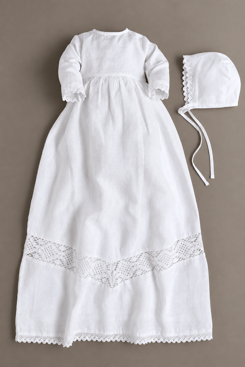 A white long-sleeve baby baptism dress with lace trim at the hem, shown flat on a taupe background alongside a matching white bonnet.