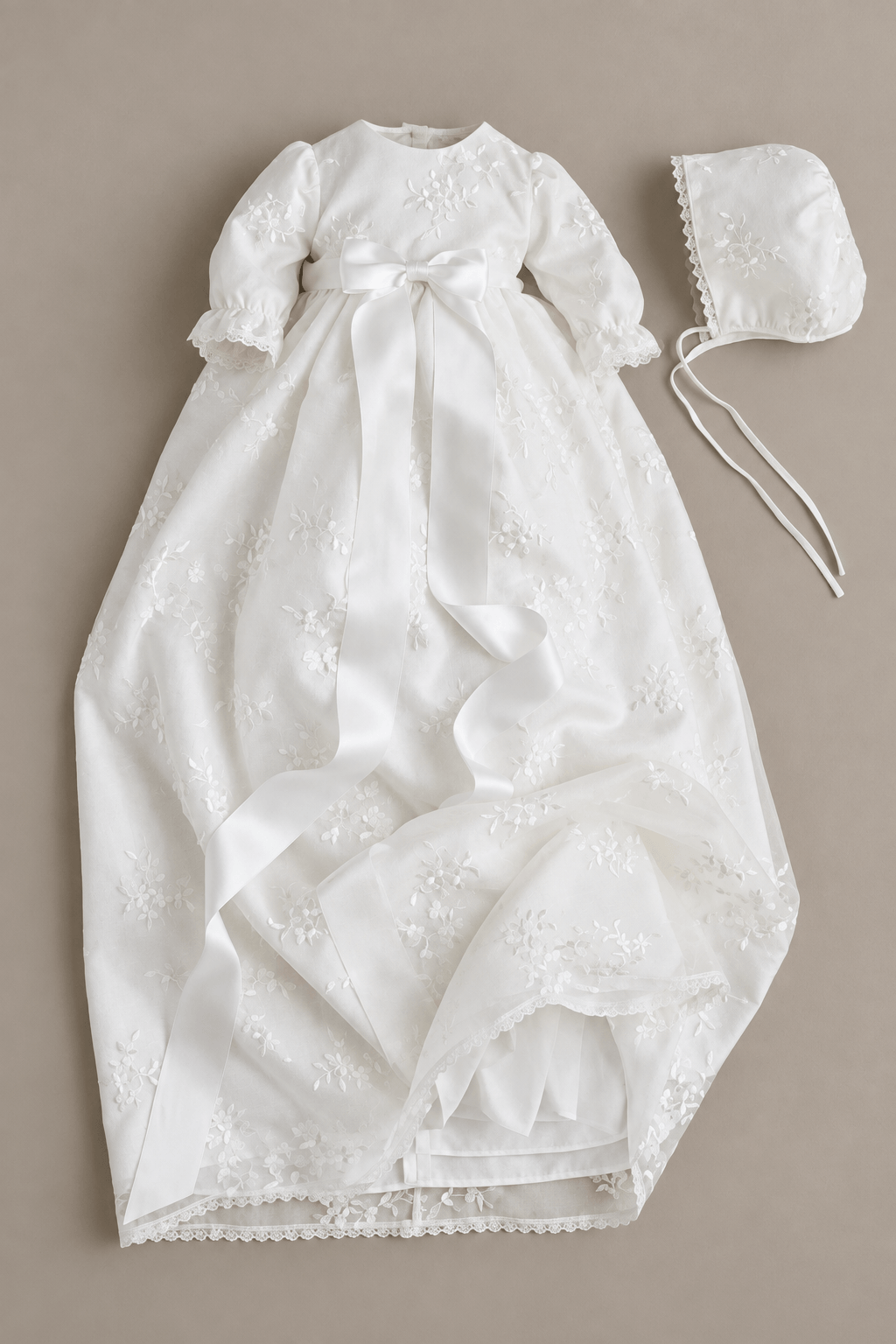 A white baby christening dress with long sleeves, a satin waist bow, and embroidered floral details, shown flat-laid next to a matching white bonnet on a neutral beige background.