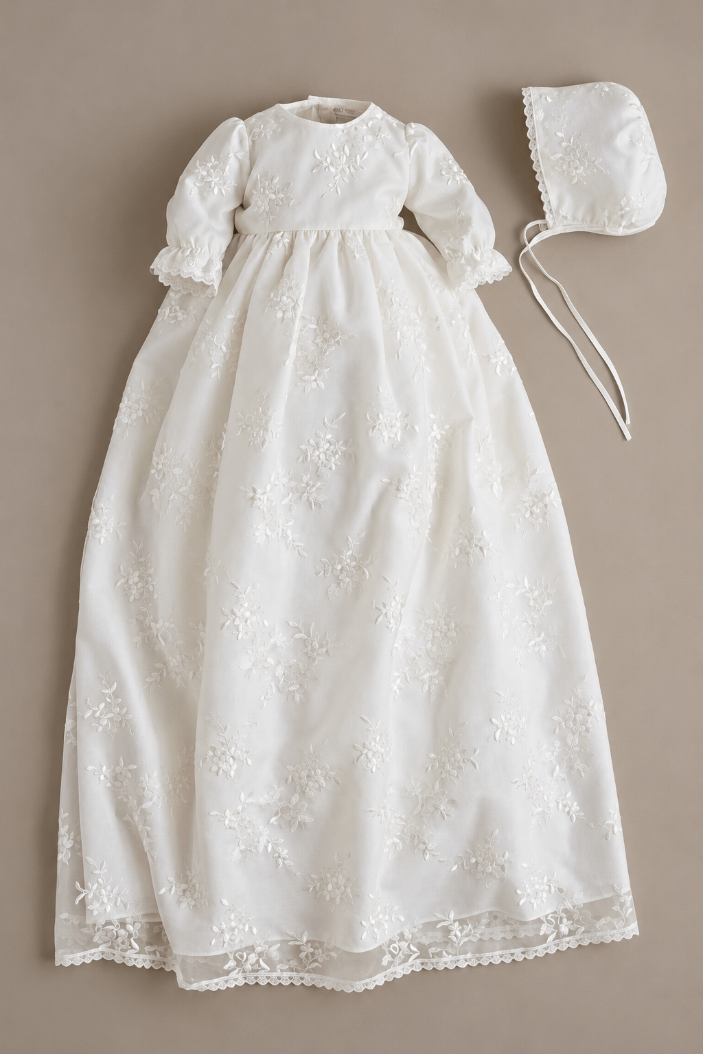 An off-white baby christening dress with embroidered floral details, shown with a matching bonnet on a solid taupe background.