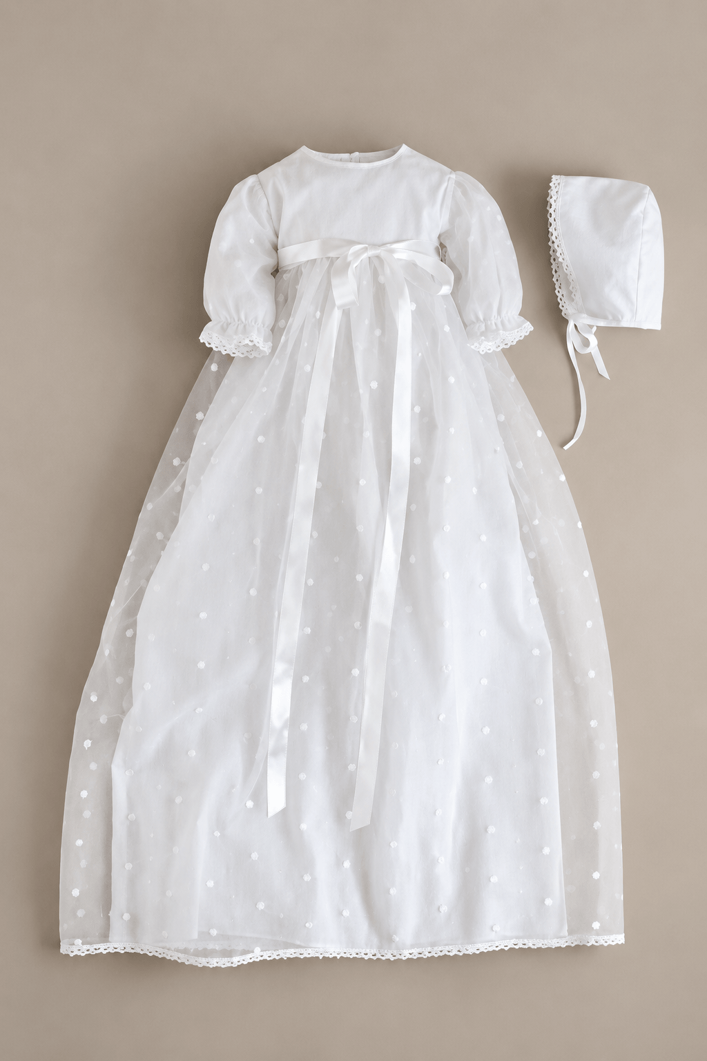 A white baby christening dress with a matching bonnet, laid flat on a neutral beige background. The dress features a white tulle skirt with tiny white embroidered dots, a white satin waist bow, and a white lace-trimmed hem.