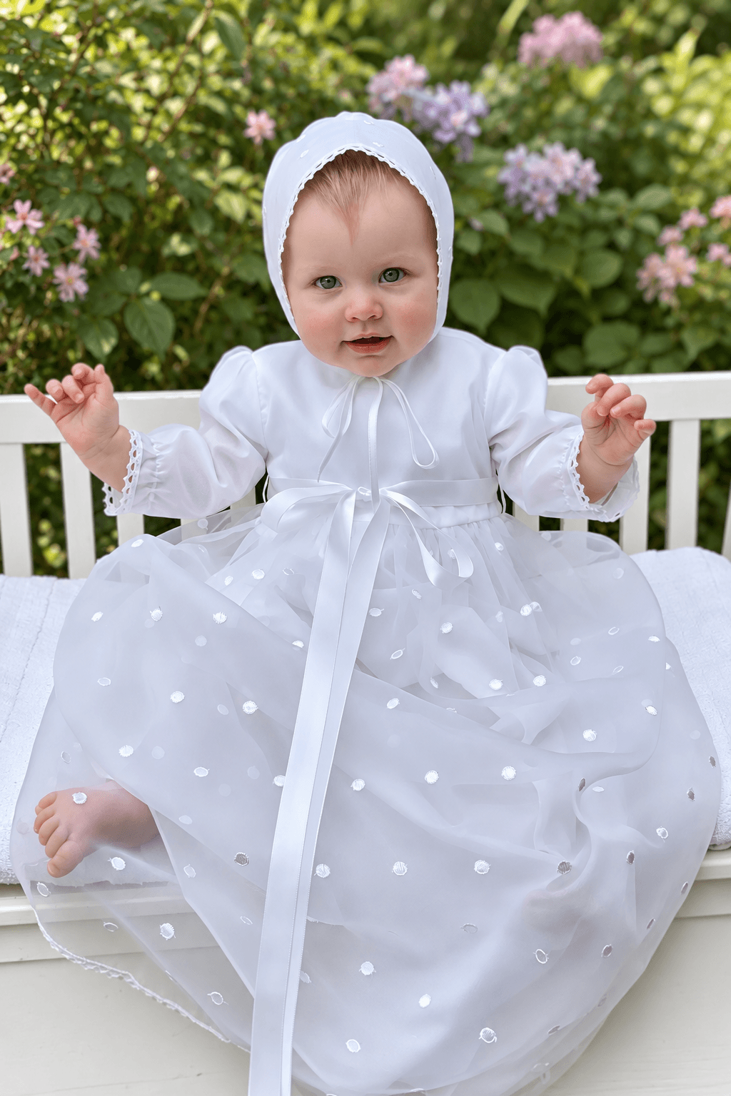 A baby wearing a white christening dress with a matching bonnet, sitting on a white bench in front of a garden background with green foliage and pink flowers.