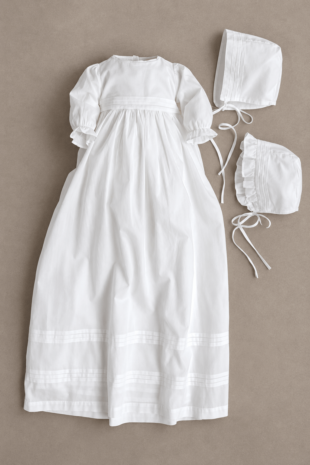 A white baby girl or boy christening dress set laid flat on a neutral taupe background, including a long smocked dress with short puff sleeves, a bonnet, and a ruffled bootie.