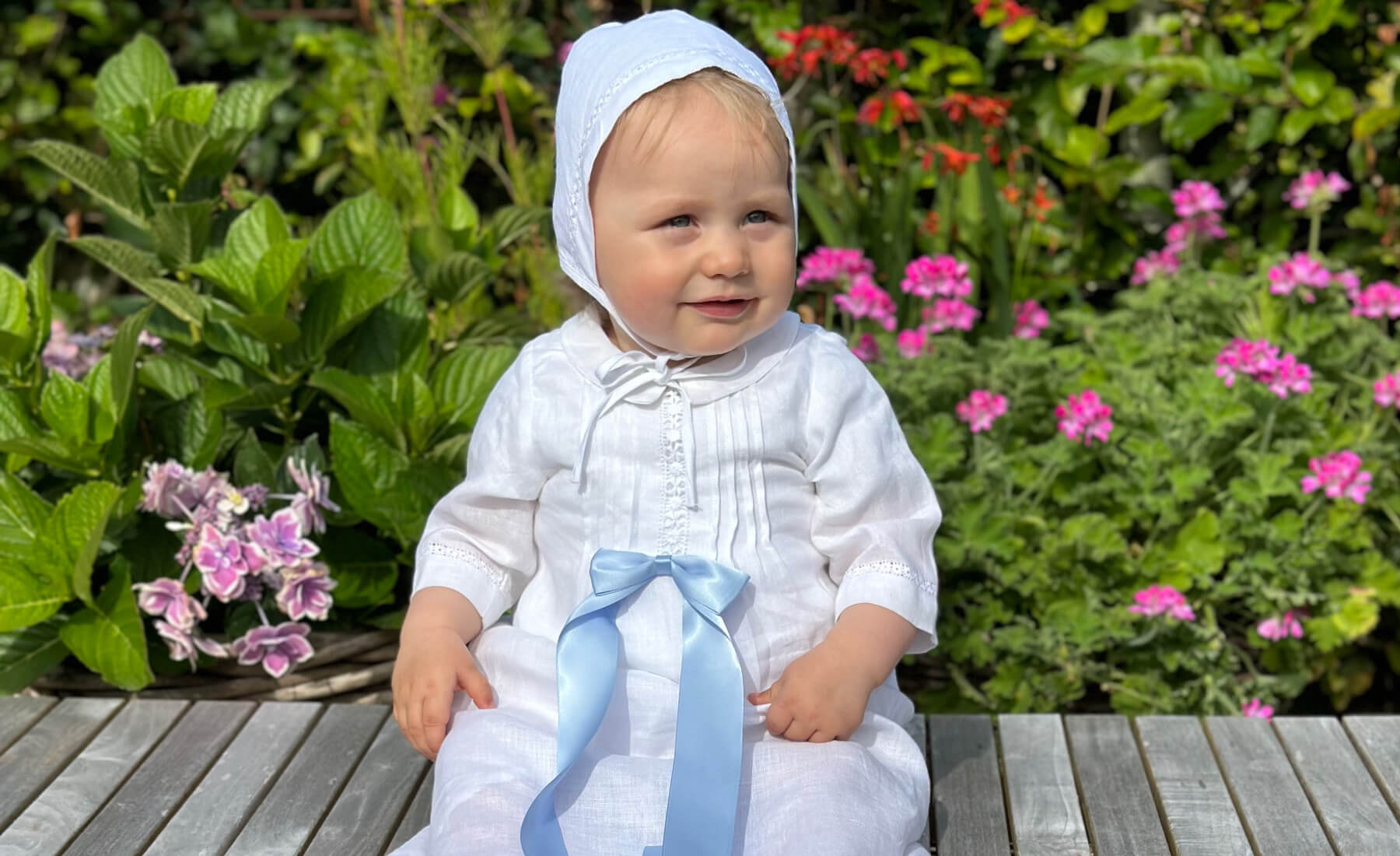Child in a white christening dress with a blue ribbon sitting on a wooden bench surrounded by flowers.
