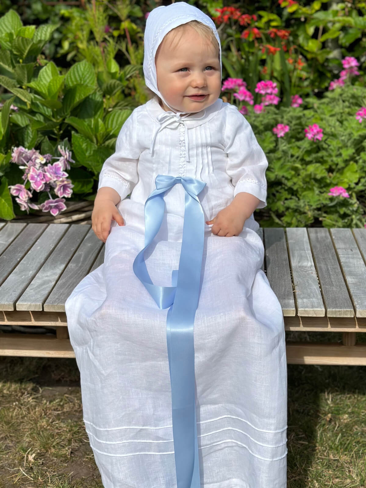 Child in a white christening dress with a blue ribbon sitting on a wooden bench surrounded by flowers.